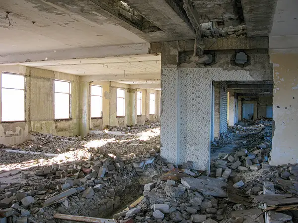 Görüntü, enkaz, kırık malzemeler ve maruz bırakılan yapısal elementlerle hasar görmüş bir iç mekan tasvir ediyor. Pencereler toz ve kirle kaplı, dışarıya bakmıyor. Tarz, gizli fotoğrafı gösteriyor.