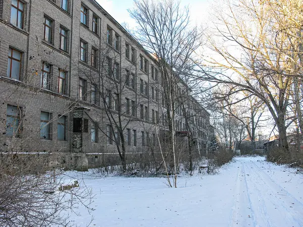 Görüntü, çok sayıda penceresi karla kaplı iki katlı eski bir tuğla bina gösteriyor. Bahçede seyrek kış bitkileri ve lastik izleri olan dar bir patika var. Gökyüzü bulutlu.