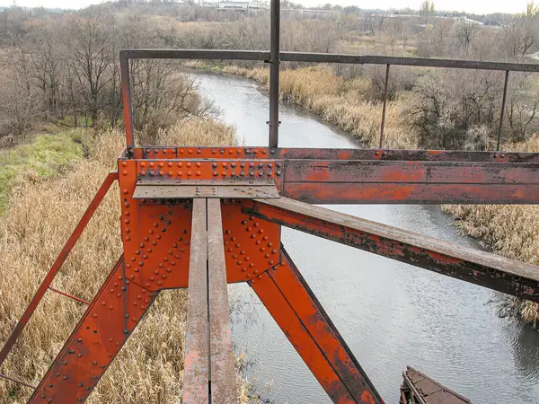 Paslı bir metal salıncak köprüsü, sonbahar ya da kış aylarında sakin ve dar bir su yolu üzerinde uzanır.