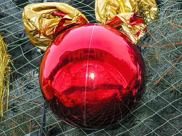 Yansıtıcı yüzeyi olan parlak kırmızı bir Noel süsü bir Noel ağacında asılı, kısmen altın sarısı bir nesne tarafından gizleniyor.