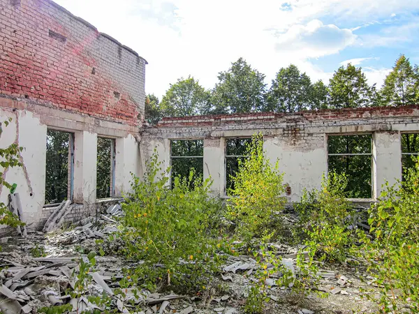 Çürümüş, terk edilmiş bir bina. Etrafı bitki örtüsü ve başka bir yapıyla çevrili. Bölge bulutlu bir gökyüzü altında.