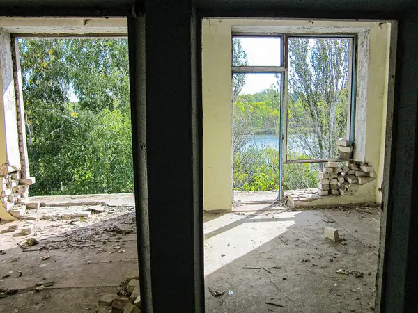Açık duvarları ve enkazı olan boş, dağınık bir oda pencerelerden geçen gün ışığı açık yeşilliği ortaya çıkarıyor, bitki örtüsü yakınındaki bir yeri gösteriyor.