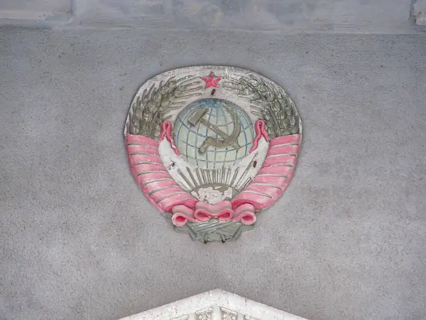 Bir çember içinde çekiç ve orak amblemi resmi, muhtemelen komünist bir rejimle ilişkili, dokulu gri bir arka planda ek dekoratif unsurlar ve mimari özellikler görünür ab