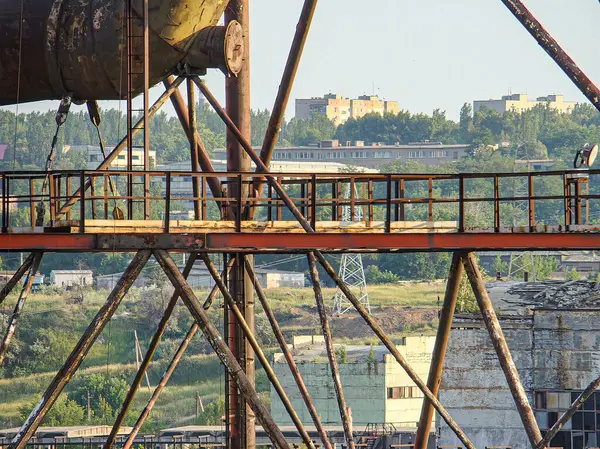 Görüntü, bitki örtüsü olan terk edilmiş bir sanayi yapısını, ön planda daha küçük yapıları, uzaktaki konut binalarını gösteriyor.