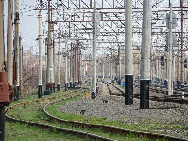 Görüntü, bulutlu bir gökyüzünün altında kırsal ya da endüstriyel bir demiryolu hattını gösteriyor. Her iki tarafında da elektrik hatları var. Raylar ve yapılar arasında çimenler, uzak bir insan ve işaretler var.