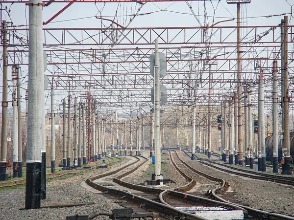 Çoklu rayları olan bir elektrikli demiryolu deposu, tepedeki katenareri yapılar, güvenlik bariyerleri ve bulutlu gökyüzünün altında trafik ışıkları