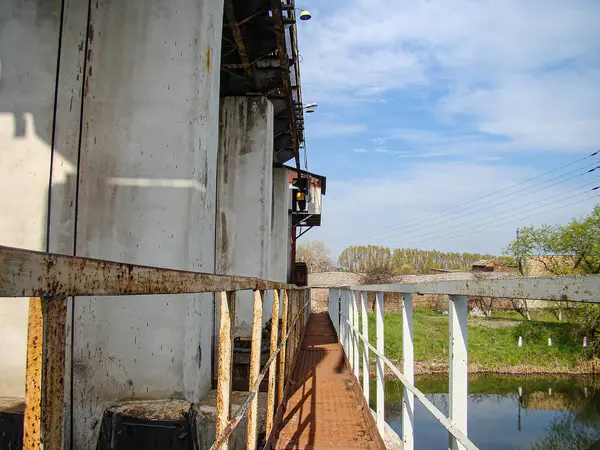 Endüstriyel bir yapının bir su kütlesi, muhtemelen bir kenetlenme alanı, ağaçlarla ve çalılarla dolu doğal bir çevrede görüntüsü. Yapı eğri büğrü metal duvarları olan bir endüstriyel binayı içeriyor.