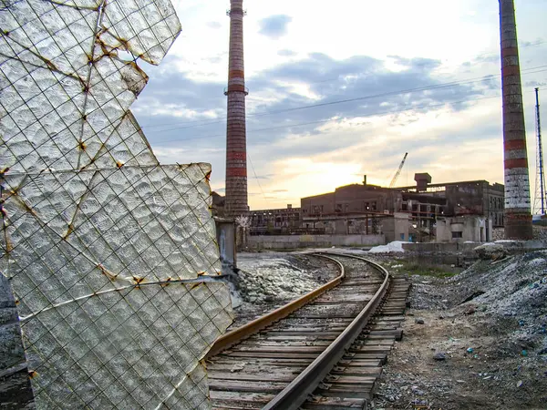 Görüntü, demiryolu rayları, enkazı ve dağınık zemini olan eski bir endüstriyel tesisi tasvir ediyor. Pist kısmen sivri cam parçalarıyla kaplı, uzun bir kuleye çıkıyor.