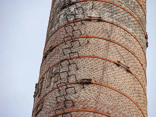 Görüntü uzun, tuğla ya da metal bacayı gösteriyor. Çoklu katmanlı ve dikey çubuklu. Gökyüzü bulutlu ve düzensiz aydınlatma, görünmeyen yapıların gölgelerini gösteriyor.