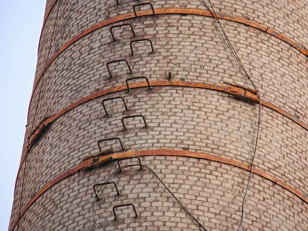 Metal basamakları ve kabloları olan uzun, tuğladan bir baca, berrak bir gökyüzüne karşı duruyor, endüstriyel mimariye odaklanıyor...