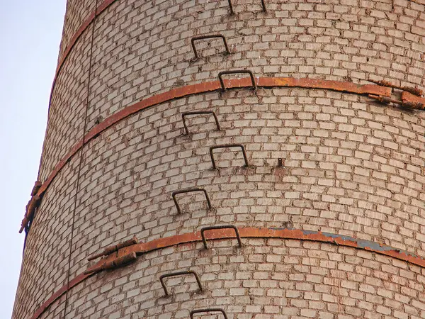 Eski, kırmızı tuğlalı bir bacaya ya da paslı metal bileşenlerine sahip bir kuleye yakın plan bulutlu bir gökyüzünü işaret ediyor.