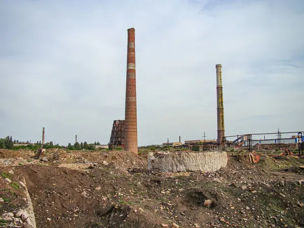 Resim, iki eski baca yığını, ön planda bir yığın atık madde, seyrek bitki örtüsü ve sisli gökyüzü ile terk edilmiş bir sanayi manzarasını tasvir ediyor.