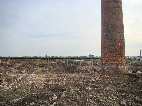 Paslı, sivri bacası olan terk edilmiş sanayi bölgesi enkaz ve molozların arasında büyüyen küçük bitkilerin kalıntıları var. Uzak yapılar, daha büyük ve karmaşık bulutlu gökyüzünü işaret ediyor.