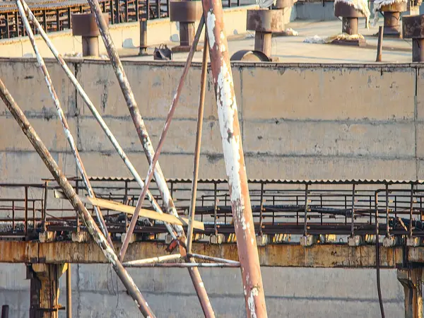 Görüntü, tersane ya da kenetlenme alanı gibi eski ve muhtemelen terk edilmiş bir sanayi ortamını gösteriyor. Yapılar yıpranmış durumda. Gri renkler bölgeye hakim ve bitki örtüsünden yeşil ipuçları geliyor.