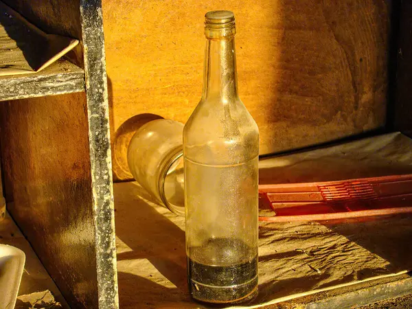Yıpranmış kenarları olan boş cam şişe, kırsal bir arka planda resmedilmiş güneş ışığı, şişeye yumuşak gölgeler ve nedenseller fırlatıyor, sıcak bir atmosfer yaratıyor. Doğal fotoğrafçılık tarzı.