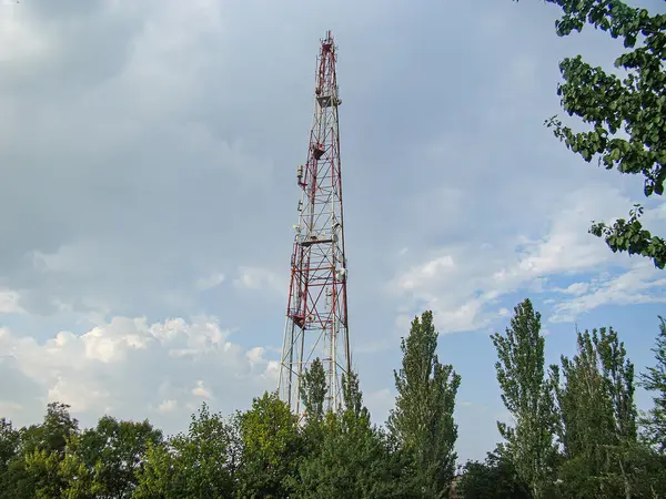 Çok katmanlı ve antenli önde gelen bir iletişim kulesinin görüntüsü. Dağınık bulutların olduğu açık bir gökyüzüne karşı duruyor. Ön plandaki ağaçların sağladığı doğal kontrastlar si için kullanılıyor.