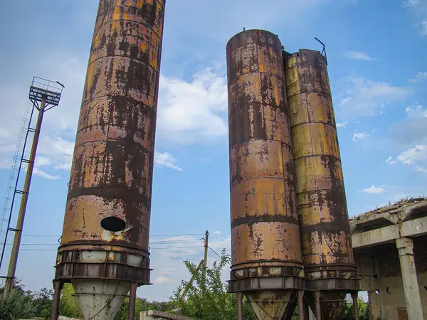 Mavi bir gökyüzünün altında iki paslı, eski silindirik tank. Elektrik hatları ve arka planda çökmekte olan bir yapı. Doğru üsteki bitki örtüsü.