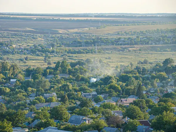 Görüntü, evleri, çatıları, yeşil yaprakları, otları, çalıları, ağaçları, eğimli araziyi, uzak evleri, temiz gökyüzünü ve hareket eden araçları gösteriyor.