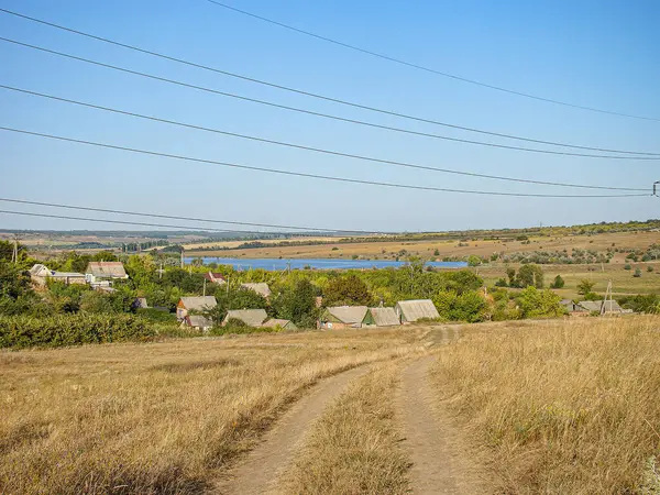 Sakin bir kırsal alan, göl kenarındaki sakin bir köye giden yıpranmış toprak bir yol, berrak mavi bir gökyüzünün altında ve elektrik hatlarıyla geçiliyor.