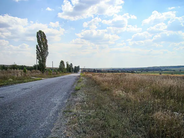 Temiz bir gökyüzünün altında yuvarlanan tepelere uzanan eski ve çatlak asfaltlı sakin bir kırsal yol sahnesi çevresi çimenli ve bitkisel.