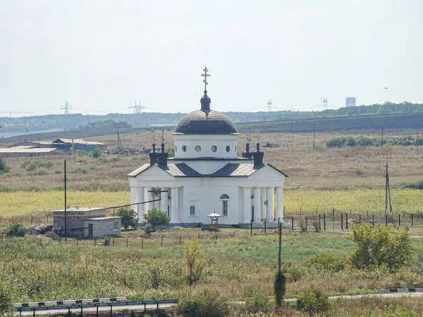 Çoklu kubbeli ve haçlı bir kırsal Doğu Ortodoks kilisesi, bulutlu gökyüzü, çimenli alanlar ve elektrik direkleriyle huzurlu bir ortamda yer alıyor. Fotoğrafın köşegen bir bakış açısı var.