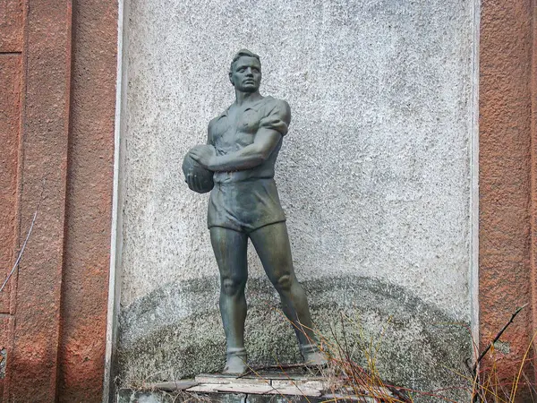 Bir top tutan orta saha oyuncusunun heykeli. Heykel çimlerin üzerinde, mimari arka planı belirsiz. Fotoğrafın gizli bir tarzı var.