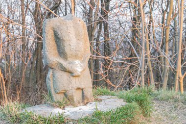 Ağaçlarla çevrili, kırsal ve aşınmış bir insan figürünü tasvir eden eski ve yıpranmış bir taş heykel.