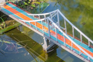 Canlı turuncu döşeme ve karmaşık parmaklıklardan oluşan minyatür beyaz ve kırmızı bir köprü, resimde resmedilmiş gibi görünüyor. Köprü suya yerleştirilmiş gibi görünüyor, bu da Ukrayna, Kyiv, Ukrayna 06-11-2024