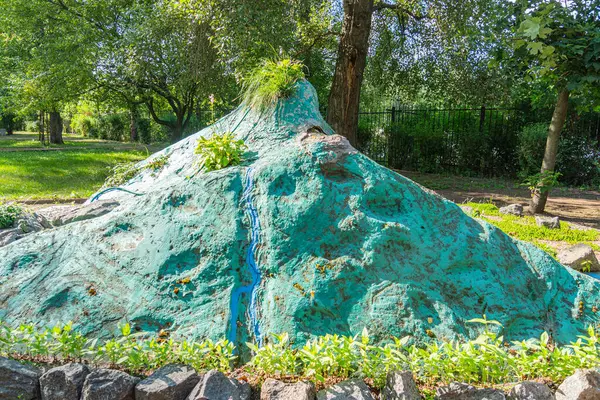 Küçük taşlarla çevrili düz bir taşın üzerinde büyük, mavi ve yeşil bir kayanın yer aldığı park veya bahçe içinde sanatsal bir kaya kurulumu. Tasarım Nat Kyiv, Ukrayna 06-11-2024 'ü birleştiriyor.