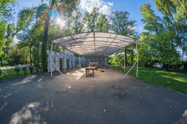 Minimalist, işlevsel bir açık hava yapısı beyaz ışıklı çatı, beton zemin, ahşap masa ve plastik sandalyeler çevrede ağaçlar ve güneş ışığı Kyiv, Ukrayna 06-11-2024