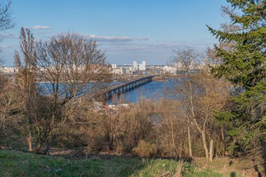Açık bir gökyüzü altında yeşil bir nehirden akan sakin bir manzara fotoğrafı, doğa ile insan gelişimi arasındaki entegrasyonu sembolize eden uzak bir köprü, Kyiv, Ukrayna, 03-29-2025
