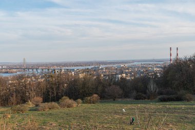 Net bir gökyüzü altında sakin, kırsal bir manzara, açık bir alanda yalnız bir figür, Kyiv, Ukrayna, 03-29-2025 kıyılarındaki uzak insan faaliyetleri ve sanayi yapılarıyla çelişiyor.