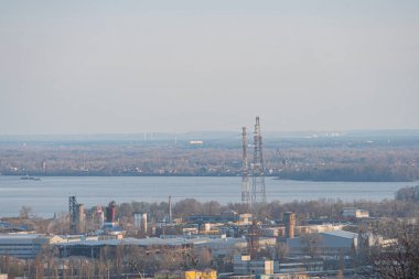 Sanayi manzarasının panoramik görüntüsü, fabrika güç santralleri, büyük bir su kütlesi ve bulanık mavi gökyüzü altında bir şehir silueti Kyiv, Ukrayna, 03-29-2025