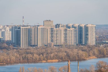 Görüntü, yüksek binalar, yansıtıcı su ve yeşillikle modern, iyi planlanmış bir şehir nehir kıyısını gösteriyor. Şehir hayatı ve doğal güzelliğin bir karışımını gösteriyor. Kyiv, Ukrayna, 03-29-2025