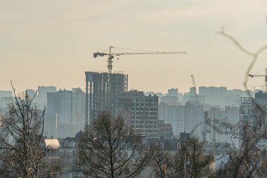 Görüntü yumuşak odaklı veya puslu bir gökyüzü altında yüksek binalarla kentsel bir ufuk çizgisini gösteriyor, bir inşaat vinci içeriyor. Bu da Kyiv, Ukrayna, 03-29-2025 kentindeki büyüme ve modernizasyonu gösteriyor.