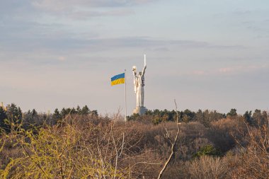 Mavi-sarı bayrağın yer aldığı bir sonbahar ya da kış manzarasına karşı bir heykel heykeli olan bulutlu bir sahne. Heykel önemli ve görüntü Kyiv, Ukrayna, 03-29-2025
