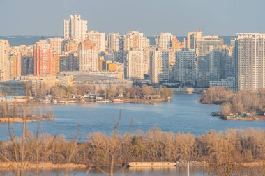 Şafak veya alacakaranlıkta canlı bir şehrin gökyüzü görüntüsü, çeşitli mimari tarzlara sahip modern yüksek binalar sergileniyor. Sahne Kyiv, Ukrayna, 03-29-2025 'i yansıtan sakin bir nehri içeriyor.