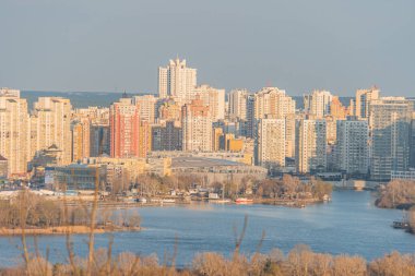 Günbatımında şehir kıyısı sahnesi, şehrin siluet çizgisinde eski ve yeni mimari tarzların karışımı sakin nehir veya körfez, orta mesafede Kyiv, Ukrayna, 03-29-2025