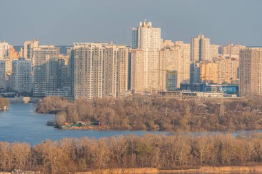 Yüksek binaları, yansıtıcı su kütlesi ve Kyiv, Ukrayna, 03-29-2025 rıhtımı boyunca yeşillikleri olan bir şehir manzarası.