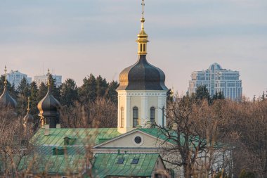 Merkezi çan kulesi, altın kulesi ve şehir merkezinde haçları olan geleneksel bir kiliseyi gösteren bir resim Ukrayna 'nın başkenti Kyiv' de, 03-29-2025 'te modern yüksek binalarda görülebilir.
