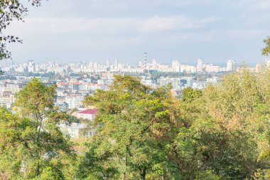 Bir tepenin tepesinden bir şehrin panoramik manzarasını gösteren çarpıcı bir görüntü, yeşil ağaçlar ve modern gökdelenlerin ahenkli bir şekilde bir arada var oluşunu gösteriyor.