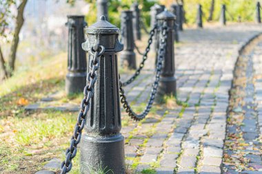 Siyah işlemeli demir kazıklar ve zincirlerle kaplı tuğla bir yol içeren davetkar, sakin bir dış mekan görüntüsü, sonbaharda bir şehir parkı ya da bahçe bölgesi olduğunu gösteriyor.