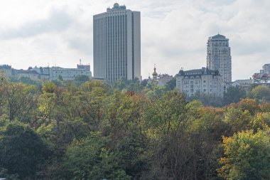 Canlı bir şehir siluetinin panoramik manzarası, yüksek görüş açısı, yüksek gökdelenler, parıldayan cam cepheler, yemyeşil, sonbahar renkleri, bulutlu bir gün, sükunet, kentsel hayat dinamizmi, harmo içindeki güzellik.