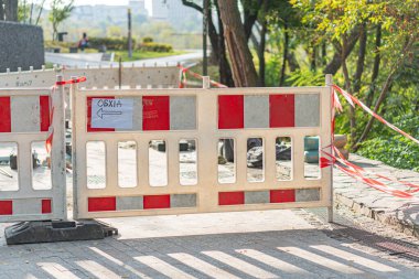 Uyarı bariyeri ve devam eden inşaat faaliyetleriyle, şehir merkezinde bir inşaat alanı tasvir ediliyor. Arka planda ağaçlar ve mavi gökyüzü yer alıyor.