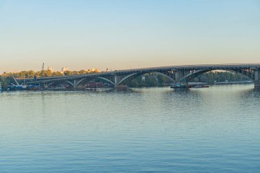 Şehir siluetinin çarpıcı panoramik görüntüsü, sakin bir nehrin üzerinde bir köprü, kentsel mimari ve doğanın karışımını yakalıyor.
