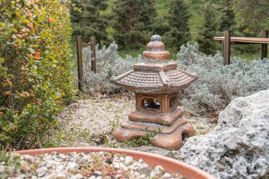 Japon mimari özelliklerine sahip eski bir taş fener, yemyeşil bitkiler ve çam ağaçlarının arasında huzurlu bir bahçede kurulmuş resim, dik bir perspektiften hafifçe yukarı bakıyor.,