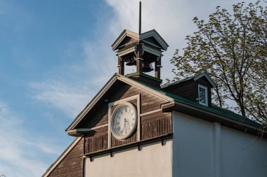 Saat kulesi olan eski ahşap kilise, açık gökyüzüne karşı ağaçların arasında duruyor. Kahverengi cephe, siyah sayılı beyaz saat, öğleden sonra sahnesi, doğal ışık, yumuşak gölgeler, güneşli gün.