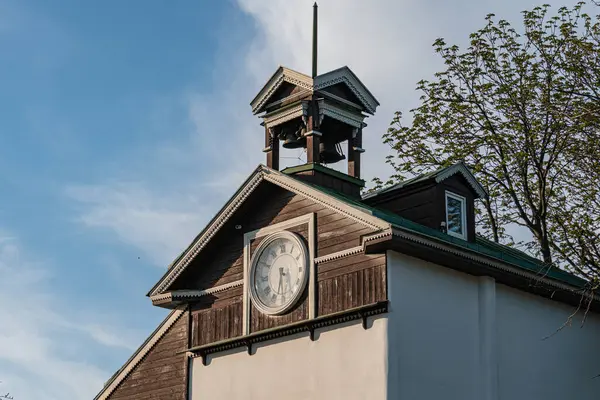 Saat kulesi olan eski ahşap kilise, açık gökyüzüne karşı ağaçların arasında duruyor. Kahverengi cephe, siyah sayılı beyaz saat, öğleden sonra sahnesi, doğal ışık, yumuşak gölgeler, güneşli gün.