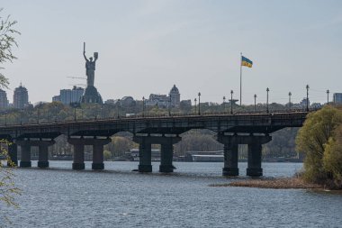 Büyük bir asma köprü, birçok çelik kule ve kabloyla desteklenen sakin bir nehri gerer. Uzakta binalar ve önemli bir anıt veya Kyiv, Ukrayna 04-17-2025