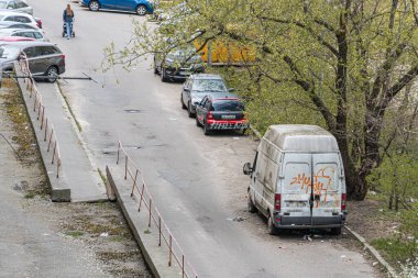 Yüksek manzaralı canlı bir şehir sokak manzarası. Sağ üst kaldırımda büyük yeşil ağaç, yürüyen kişi, park etmiş arabalar, turuncu yazılı beyaz minibüs, Mov Kyiv, Ukrayna 04-19-2025.